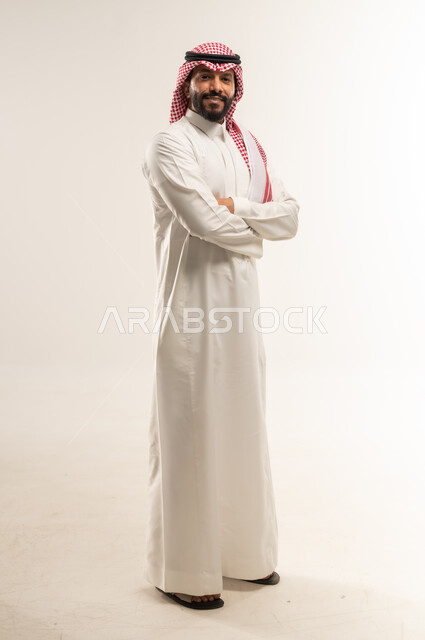 الاهتمام بالأناقة والمظهر الخارجي، النظر إلى الكاميرا بايماءات الجدية والسرور، مفهوم الرجولة والقوة والثقة بالنفس، بورتريه لرجل عربي خليجي بحريني يرتدي الثوب التقليدي والشماغ يقف بأيدي متقاطعة بتعابير الحزم، الطول الكامل للجسم، خلفية بيضاء