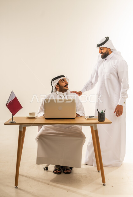 التحاور وتبادل الخبرات بين الموظفين، مفهوم العمل الجماعي وروح الفريق، إبداء الرأي حول استراتيجات وأهداف الشركة، بورتريه لرجل عربي خليجي قطري يرتدي الثوب التقليدي والغترة يجلس خلف طاولة المكتب وبجانبه علم قطر يتناقش مع زميله حول أمور العمل، خلفية بيضاء