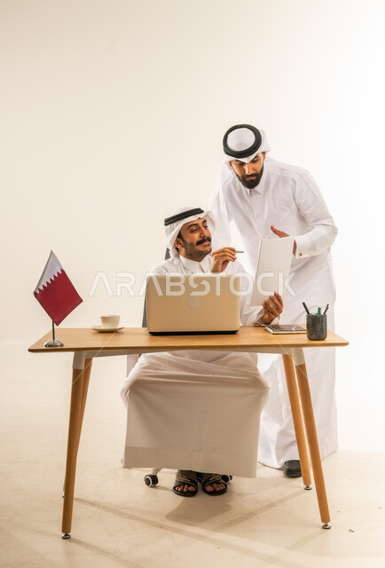 مفهوم العمل الجماعي وروح الفريق، التحاور وتبادل الخبرات بين الموظفين، إبداء الرأي حول استراتيجات وأهداف الشركة، بورتريه لرجل عربي خليجي قطري يرتدي الثوب التقليدي والغترة يجلس خلف طاولة المكتب وبجانبه علم قطر يتناقش مع زميله حول أمور العمل، خلفية بيضاء
