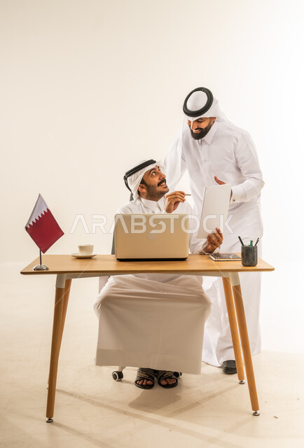 إبداء الرأي حول الاستراتيجات والأهداف، التحاور وتبادل الخبرات بين الموظفين، استخدام الحاسوب المحمول بالعمل، بورتريه لرجل عربي خليجي قطري يرتدي الثوب التقليدي والغترة يجلس خلف طاولة المكتب وبجانبه علم قطر يتناقش مع زميله حول أمور العمل، خلفية بيضاء