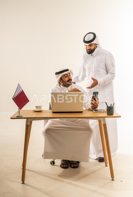 تبادل الخبرات والمهارات بين الزملاء، إنجاز المهام باستخدام الحاسوب المحمول، دمج التقنية والتكنولوجيا بالمجال المكتبي، بورتريه لرجل عربي خليجي قطري يرتدي الثوب التقليدي والغترة يجلس خلف المكتب بجانبه علم قطر ويمسك الجوال بيده يتحدث مع زميله بالعمل، خلفية بيضاء