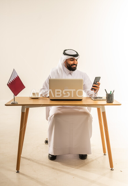 استخدام الهاتف المحمول في العمل، إقامة الاجتماعات أونلاين عبر مكالمات الفيديو، الرد والإجابة على الأسئلة والاستفسارات والمكالمات، بورتريه لرجل عربي خليجي قطري يرتدي الثوب التقليدي والغترة يجلس خلف المكتب أمامه الحاسوب المحمول وعلم قطر، خلفية بيضاء