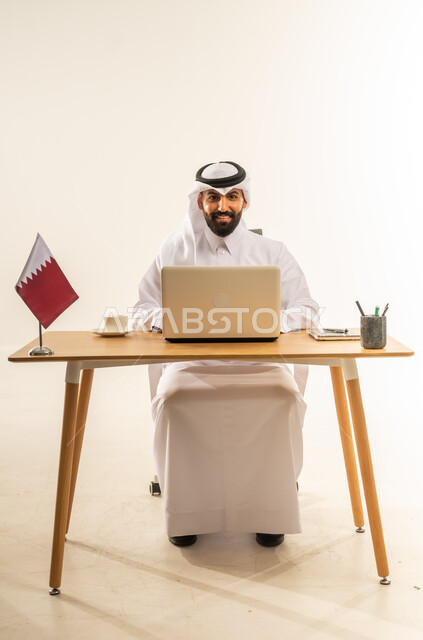 دمج التقنيات الحديثة بالمجال الإداري، العمل في المكاتب والشركات القطرية، الاستمتاع بتناول فنجان من القهوة الساخنة، بورتريه لرجل عربي خليجي قطري يرتدي الثوب التقليدي والغترة يجلس خلف طاولة المكتب وبجانبه علم قطر يعمل على الحاسوب المحمول، خلفية بيضاء
