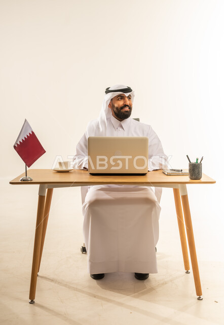 العمل في المكاتب والشركات القطرية، الاستمتاع بتناول فنجان من القهوة الساخنة، دمج التقنيات الحديثة بالمجال الإداري، بورتريه لرجل عربي خليجي قطري يرتدي الثوب التقليدي والغترة يجلس خلف طاولة المكتب وبجانبه علم قطر يعمل على الحاسوب المحمول، خلفية بيضاء