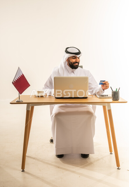 عملية الدفع الالكتروني بواسطة بطاقة الائتمان، مفهوم التسوق أونلاين، الشراء عبر تطبيقات ومواقع الانترنت، التبضع عن بعد بسهولة ويسر، بورتريه لرجل عربي خليجي قطري يرتدي الثوب التقليدي والغترة يجلس خلف المكتب أمامه الحاسوب المحمول وعلم قطر، خلفية بيضاء