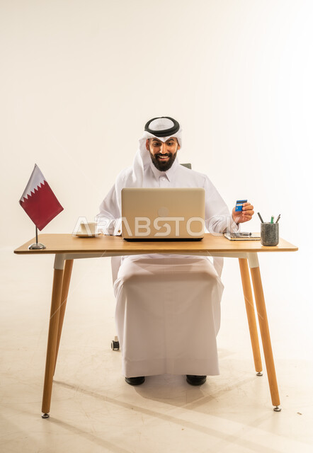 مفهوم التسوق أونلاين، عملية الدفع الالكتروني بواسطة بطاقة الائتمان، الشراء عبر تطبيقات ومواقع الانترنت، التبضع عن بعد بسهولة ويسر، بورتريه لرجل عربي خليجي قطري يرتدي الثوب التقليدي والغترة يجلس خلف المكتب أمامه الحاسوب المحمول وعلم قطر، خلفية بيضاء