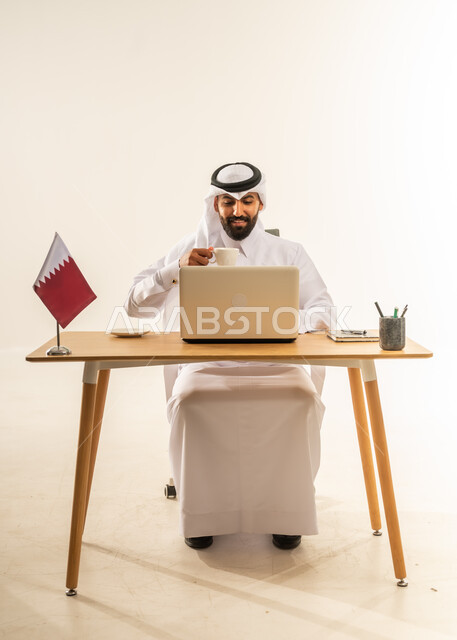 الاستمتاع بتناول فنجان من القهوة الساخنة، دمج التقنيات الحديثة بالمجال الإداري، العمل في المكاتب والشركات القطرية، بورتريخ لرجل عربي خليجي قطري يرتدي الثوب التقليدي والغترة يجلس خلف طاولة المكتب وبجانبه علم قطر يعمل على الحاسوب المحمول، خلفية بيضاء