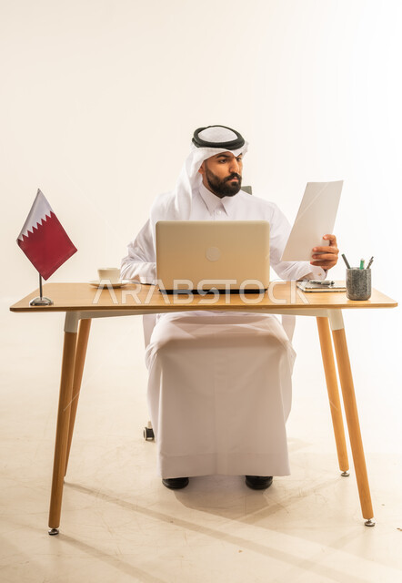 إنجاز المهام باحترافية باستخدام الحاسوب المحمول، دمج التكنولوجيا والتقنيات الحديثة في المجال الإداري، العمل في مكاتب قطر، بورتريه لرجل عربي خليجي قطري يرتدي الثوب التقليدي والغترة يجلس خلف طاولة المكتب وبجانبه علم قطر يمسك الأوراق بيده، خلفية بيضاء