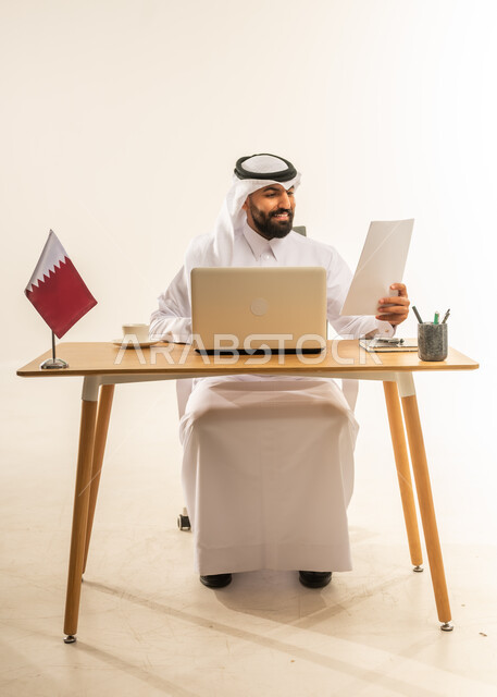 دمج التكنولوجيا والتقنيات الحديثة في المجال الإداري، إنجاز المهام باحترافية باستخدام الحاسوب المحمول، العمل في مكاتب قطر، بورتريه لرجل عربي خليجي قطري يرتدي الثوب التقليدي والغترة يجلس خلف طاولة المكتب وبجانبه علم قطر يمسك الأوراق بيده، خلفية بيضاء