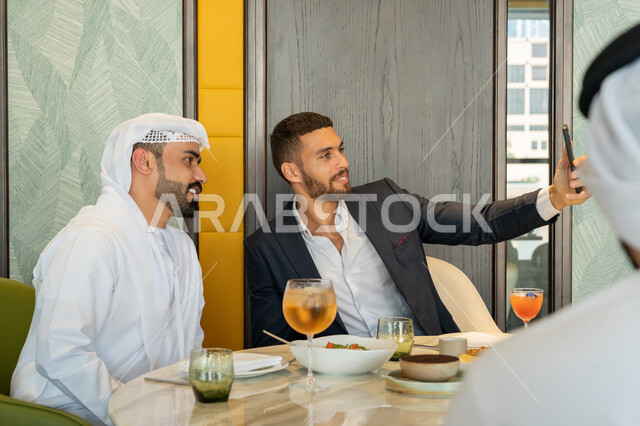 توثيق اللحظات السعيدة, قضاء الأوقات الممتعة مع الأصحاب, استخدام الأجهزة التقنية الحديثة، أصدقاء عربيين خليجيين إماراتيين يجلسون على المائدة بإحدى المقاهي ويقومون بالتقاط الصور التذكارية عبر الجوال