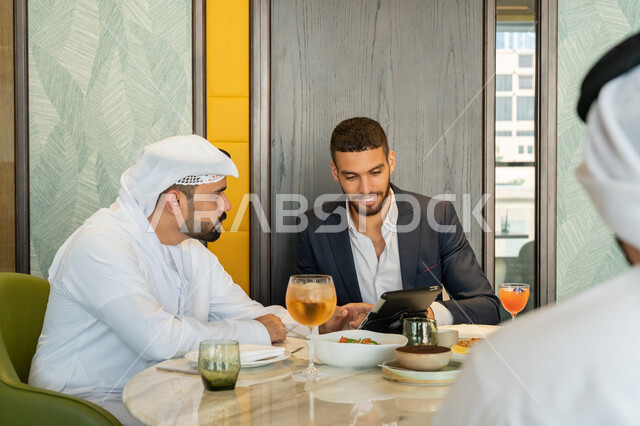 Using modern technical devices, dialogues and discussions between colleagues, a group of Arab Gulf Emirati friends sitting at a table in a restaurant in the Emirates and using a tablet, spending enjoyable times in the restaurant