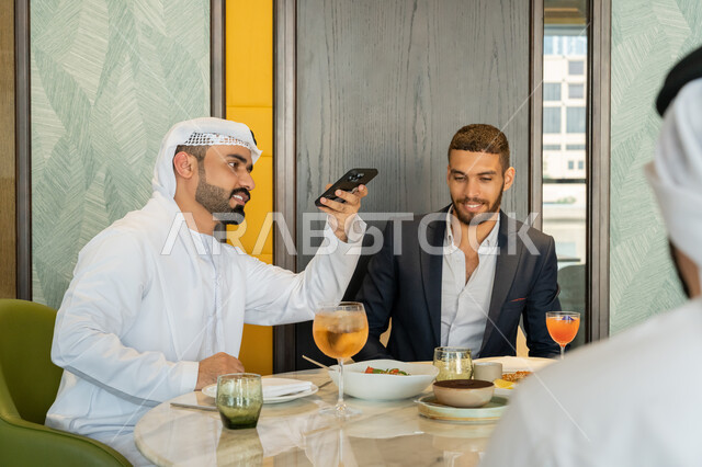 توثيق اللحظات السعيدة, قضاء الأوقات الممتعة مع الأصحاب, استخدام الأجهزة التقنية الحديثة، أصدقاء عربيين خليجيين إماراتيين يجلسون على المائدة بإحدى المقاهي ويقومون بالتقاط الصور التذكارية عبر الجوال