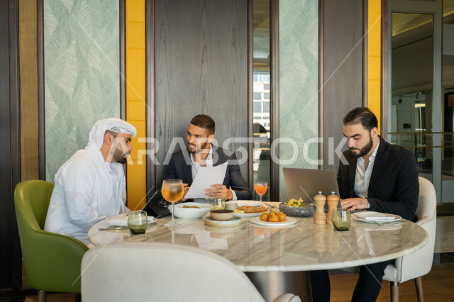 استخدام الأجهزة التقنية الحديثة, الحوارات والنقاشات بين الزملاء, استعمال جهاز الحاسوب المحمول, مناقشة مهام العمل في المطعم, مجموعة من الأصدقاء العربيين الخليجيين السعوديين يجلسون على المائدة في إحدى المطاعم في الإمارات