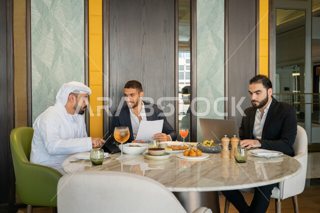 الحوارات والنقاشات بين الزملاء, استخدام الأجهزة التقنية الحديثة, استعمال جهاز الحاسوب المحمول, مناقشة مهام العمل في المطعم, مجموعة من الأصدقاء العربيين الخليجيين السعوديين يجلسون على المائدة في إحدى المطاعم في الإمارات