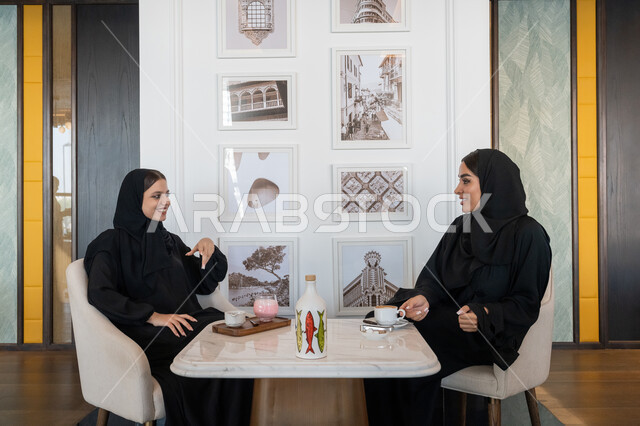 بناء علاقات اجتماعية ناجحة, النقاشات والحوارات في الأجواء الهادئة, الاستمتاع بالمشروبات الباردة، امرأة عربية خليجية إماراتية محجبة ترتدي العباءة السوداء تجلس مع صديقتها بإحدى المقاهي, شابتان سعوديتان تقضيان أوقات ممتعة ومسلية