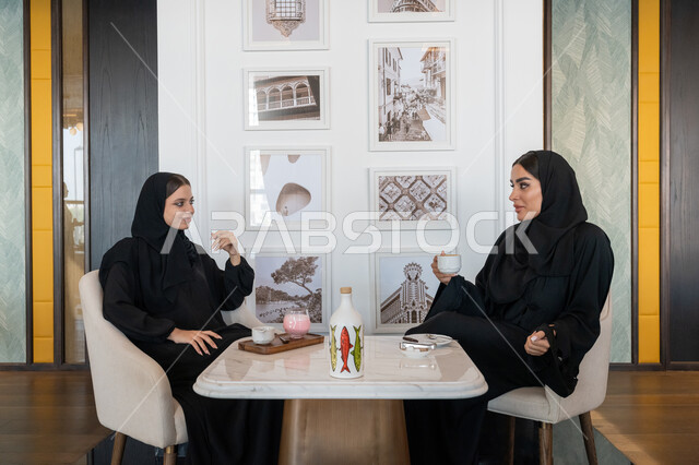الاستمتاع بالمشروبات الباردة, النقاشات والحوارات في الأجواء الهادئة، بناء علاقات اجتماعية ناجحة، امرأة عربية خليجية إماراتية محجبة ترتدي العباءة السوداء تجلس مع صديقتها بإحدى المقاهي, شابتان سعوديتان تقضيان أوقات ممتعة ومسلية