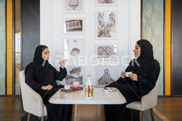 Having fun and entertaining times with friends, building successful social relationships, discussions and dialogues in a calm atmosphere, a veiled Emirati Gulf Arab woman wearing a black abaya sitting with her friend in a cafe, two young Saudi women enjoying cold drinks