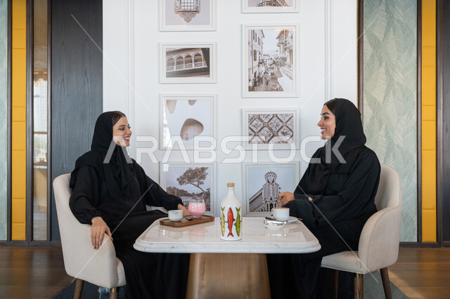 بناء علاقات اجتماعية ناجحة, النقاشات والحوارات في الأجواء الهادئة, الاستمتاع بالمشروبات الباردة، امرأة عربية خليجية إماراتية محجبة ترتدي العباءة السوداء تجلس مع صديقتها بإحدى المقاهي, شابتان سعوديتان تقضيان أوقات ممتعة ومسلية