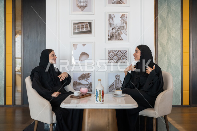 الاستمتاع بالمشروبات الباردة, النقاشات والحوارات في الأجواء الهادئة، بناء علاقات اجتماعية ناجحة، امرأة عربية خليجية إماراتية محجبة ترتدي العباءة السوداء تجلس مع صديقتها بإحدى المقاهي, شابتان سعوديتان تقضيان أوقات ممتعة ومسلية