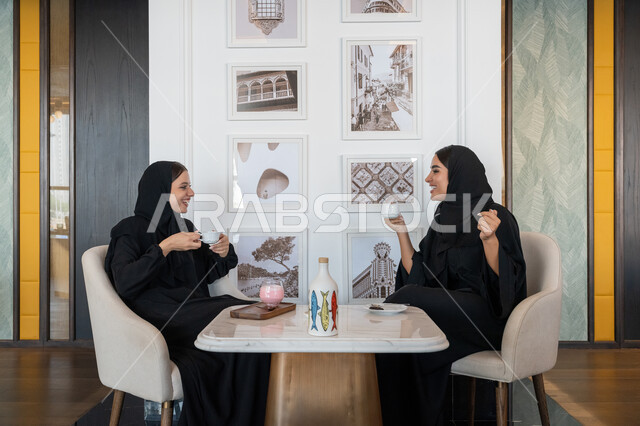بناء علاقات اجتماعية ناجحة, النقاشات والحوارات في الأجواء الهادئة, الاستمتاع بالمشروبات الباردة، امرأة عربية خليجية إماراتية محجبة ترتدي العباءة السوداء تجلس مع صديقتها بإحدى المقاهي, شابتان سعوديتان تقضيان أوقات ممتعة ومسلية
