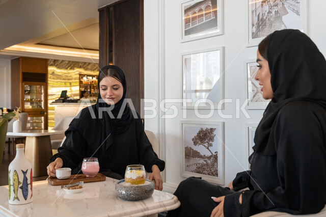 النقاشات والحوارات في الأجواء الهادئة، بناء علاقات اجتماعية ناجحة، الاستمتاع بالمشروبات الباردة، امرأة عربية خليجية إماراتية محجبة ترتدي العباءة السوداء تجلس مع صديقتها بإحدى المقاهي, شابتان سعوديتان تقضيان أوقات ممتعة ومسلية