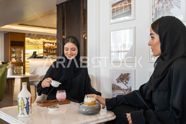 الاستمتاع بالمشروبات الباردة, النقاشات والحوارات في الأجواء الهادئة، بناء علاقات اجتماعية ناجحة، امرأة عربية خليجية إماراتية محجبة ترتدي العباءة السوداء تجلس مع صديقتها بإحدى المقاهي, شابتان سعوديتان تقضيان أوقات ممتعة ومسلية