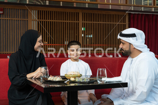 الأجواء العائلية السعيدة, مطاعم وكافيهات الإمارات, مشاركة الأحاديث والأخبار مع العائلة, قضاء الأوقات الممتعة في المطعم, عائلة عربية خليجية إماراتية يرتدون الثوب الإماراتي التقليدي يجلسون على المائدة بتعابير السعادة والسرور