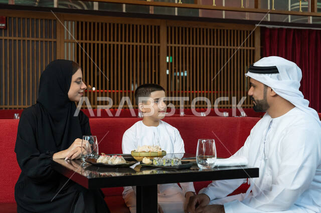 مشاركة الأحاديث والأخبار مع العائلة, الأجواء العائلية السعيدة, مطاعم وكافيهات الإمارات, قضاء الأوقات الممتعة في المطعم, عائلة عربية خليجية إماراتية يرتدون الثوب الإماراتي التقليدي يجلسون على المائدة بتعابير السعادة والسرور