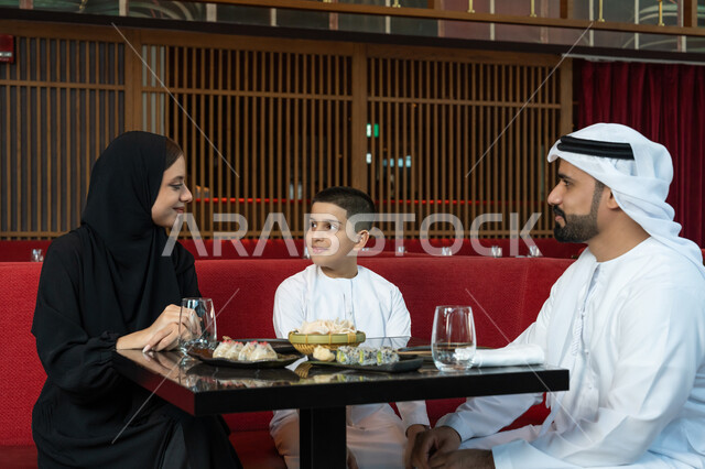 الأجواء العائلية السعيدة, مطاعم وكافيهات الإمارات, مشاركة الأحاديث والأخبار مع العائلة, قضاء الأوقات الممتعة في المطعم, عائلة عربية خليجية إماراتية يرتدون الثوب الإماراتي التقليدي يجلسون على المائدة بتعابير السعادة والسرور
