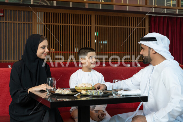 مشاركة الأحاديث والأخبار مع العائلة, الأجواء العائلية السعيدة, مطاعم وكافيهات الإمارات, قضاء الأوقات الممتعة في المطعم, عائلة عربية خليجية إماراتية يرتدون الثوب الإماراتي التقليدي يجلسون على المائدة بتعابير السعادة والسرور