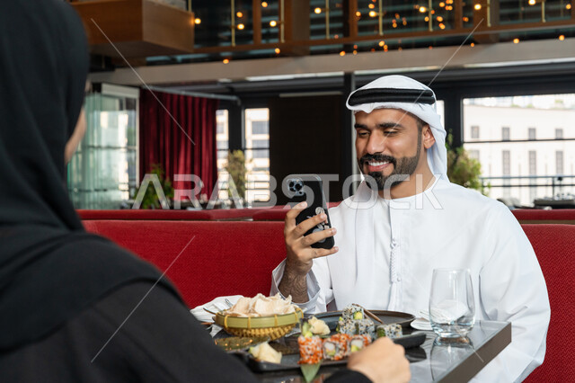 Spending fun family time, using modern technology devices, an Emirati Gulf Arab man wearing a white kandura and ghutra sitting at the dining table with his wife and using a mobile phone, restaurants and cafes in the United Arab Emirates