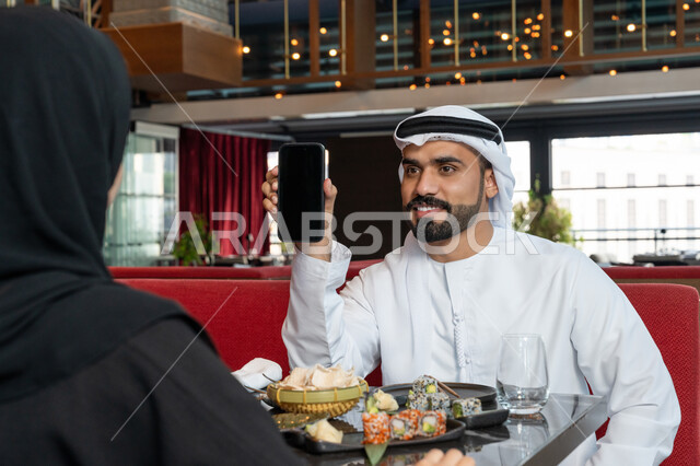 استخدام الأجهزة التقنية الحديثة, قضاء الأوقات العائلية الممتعة, رجل عربي خليجي إماراتي يرتدي الكندورة و الغترة البيضاء يجلس على مائدة الطعام مع زوجته ويمسك بيده الهاتف المحمول, مطاعم و كافيهات في الإمارات العربية المتحدة, عرض شاشة سوداء فارغة عبر الجوال