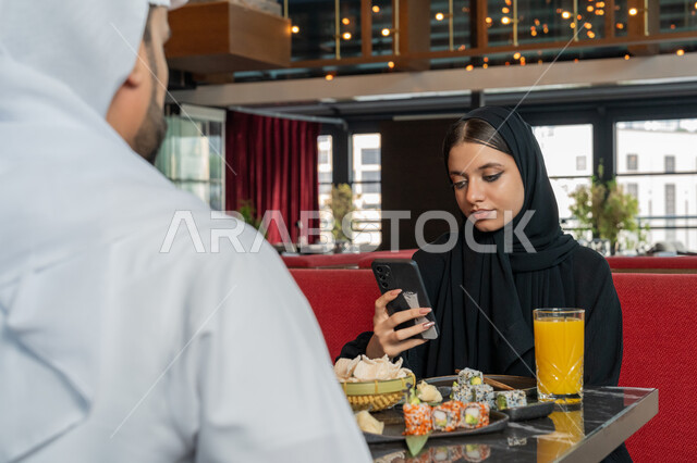 استخدام الأجهزة التقنية الحديثة, قضاء الأوقات الممتعة في المطعم, امرأة عربية خليجية إماراتية ترتدي الزي التقليدي الإماراتي تجلس على المائدة مع زوجها و تقوم باستخدام الهاتف المحمول, مطاعم و كافيهات في الإمارات العربية المتحدة