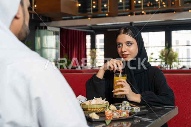 الاستمتاع بالمشروبات الباردة, قضاء الأوقات الممتعة في المطعم, امرأة عربية خليجية إماراتية محجبة ترتدي العباءة السوداء تجلس على المائدة مع زوجها و تقوم بشرب العصير الطازج, مطاعم و كافيهات في الإمارات العربية المتحدة
