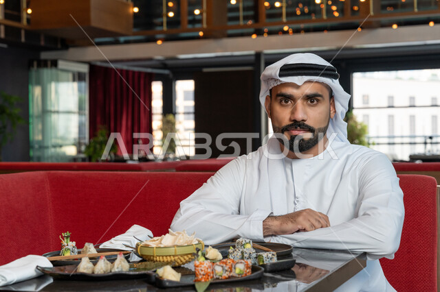 مطاعم و كافيهات في الإمارات العربية المتحدة, قضاء الأوقات الممتعة في المطعم, رجل عربي خليجي إماراتي يرتدي الكندورة والغترة البيضاء يجلس على المائدة في المطعم وينظر بإيماءات السعادة والسرور, تشكيلة متنوعة من المأكولات