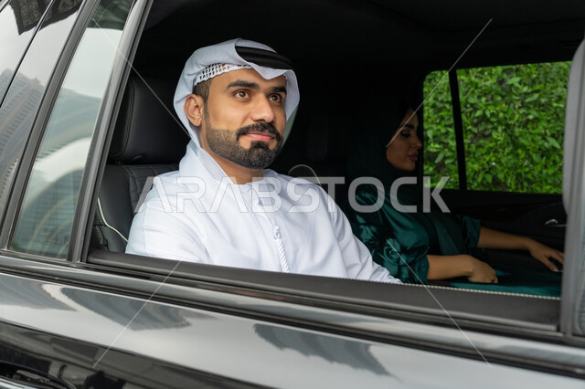 استخدام التاكسي للوصول إلى الوجهة المطلوبة, خدمة توصيل الركاب، خدمات النقل الآمنة في الإمارات العربية المتحدة، صورة مقربة لرجل عربي خليجي إماراتي يرتدي الكندورة والغترة البيضاء يجلس داخل السيارة مع زوجته وينظر بإيماءات السعادة والسرور