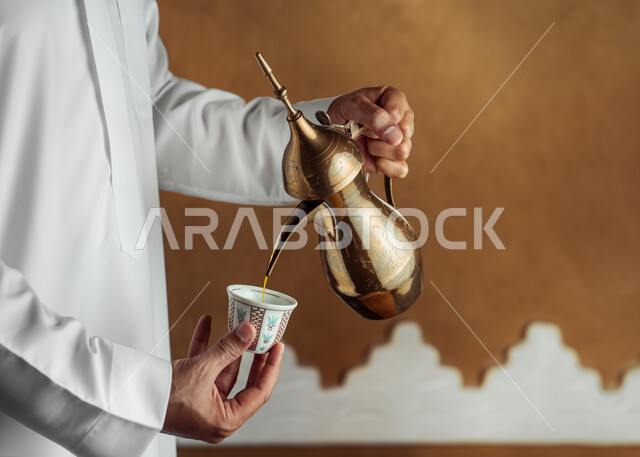 The concept of generosity and hospitality, the anniversary of the founding of the first Saudi state, February 22, the day of our beginning 1727 AD, celebrating national occasions and holidays, a close-up of a Saudi Gulf Arab man wearing traditional costumes pouring coffee from a copper pot, offering local products to guests