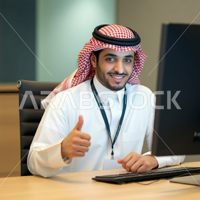 التعبير عن الرأي بالاعجاب والقبول, صورة مقربة لرجل عربي خليجي سعودي يرتدي الثوب والشماغ التقليدي يجلس في مكتبه ويقوم برفع اصبع الابهام للأعلى بإيماءات الامتياز و علامة الجودة, النظر إلى الكاميرا بإشارات الموافقه