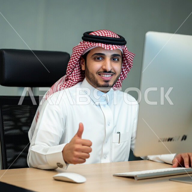 التعبير عن الرأي بالاعجاب والقبول, صورة مقربة لرجل عربي خليجي سعودي يرتدي الثوب والشماغ التقليدي يجلس في مكتبه ويقوم برفع اصبع الابهام للأعلى بإيماءات الامتياز و علامة الجودة, النظر إلى الكاميرا بإشارات الموافقه