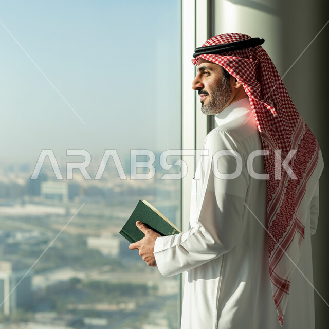 Enjoying reading books, office administrative professions and jobs, managing and organizing business affairs, a picture from behind of a Saudi Arabian Gulf businessman wearing traditional clothing and a shemagh, standing and holding a book in his hand, practicing the hobby of reading