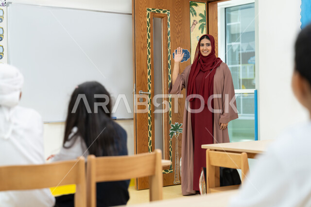 أهداف التربية والتعليم بالمناهج المقررة للطلاب والطالبات في الإمارات العربية المتحدة، معلمة عربية خليجية إماراتية ترتدي الحجاب والعباءة ترفع يدها للأعلى ترحب بالتلاميذ بإيماءات السرور، الترحيب وإلقاء التحية، المشاركة والتفاعل داخل الفصل الدراسي