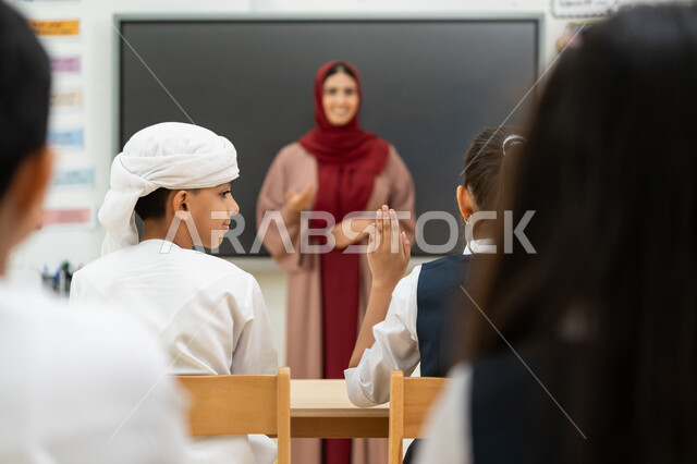 طالبة ترفع يدها للإجابة على أسئلة المدرسة، أهداف التربية والتعليم بالمناهج المقررة للطلاب والطالبات في الإمارات العربية المتحدة، معلمة عربية خليجية إماراتية ترتدي الحجاب والعباءة تجلس خلف مكتبها، لوح مدرسي ذكي، المشاركة والتفاعل داخل الفصل الدراسي