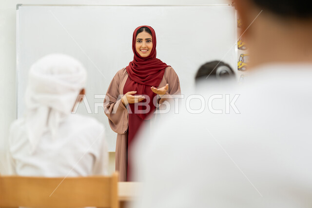 لوحة مدرسية بيضاء، المشاركة داخل الفصل الدراسي، معلمة عربية خليجية إماراتية ترتدي الحجاب والعباءة ترفع يديها تقوم بشرح الدروس للتلاميذ، أهداف التربية والتعليم بالمناهج المقررة للطلاب والطالبات في الإمارات العربية المتحدة