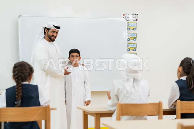 التفاعل والنشاط داخل الفصل الدراسي، لوحة مدرسية بيضاء، معلم عربي خليجي إماراتي يرتدي الكندورة والغترة يرحب بطالب جديد بإيماءات السرور، تعريف الطلاب على زميلهم الجديد، أهداف التربية والتعليم بالمناهج المقررة للطلاب والطالبات في الإمارات العربية المتحدة