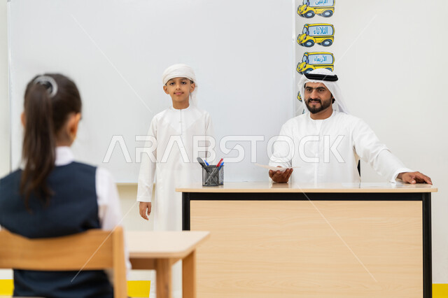 طالب يقف أمام لوحة مدرسية بيضاء يشرح الدرس، التفاعل والنشاط داخل الفصل الدراسي، معلم عربي خليجي إماراتي يرتدي الكندورة والغترة يجلس خلف مكتبه، أهداف التربية والتعليم بالمناهج المقررة للطلاب والطالبات في الإمارات العربية المتحدة