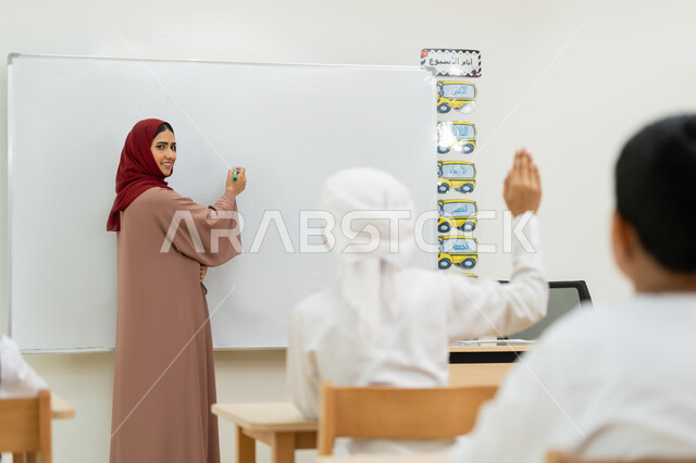 طالب يرفع يده للإجابة على أسئلة المدرسة، معلمة عربية خليجية إماراتية ترتدي الحجاب والعباءة تقف أمام اللوحة المدرسية البيضاء تشرح الدرس، التفاعل والنشاط داخل الفصل الدراسي، أهداف التربية والتعليم بالمناهج المقررة للطلاب والطالبات في الإمارات العربية المتحدة