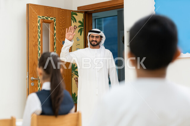 الترحيب وإلقاء التحية، التفاعل والنشاط داخل الفصل الدراسي، معلم عربي خليجي إماراتي يرتدي الكندورة والغترة يدخل الصف ويرفع يده مرحباً بالتلاميذ، أهداف التربية والتعليم بالمناهج المقررة للطلاب والطالبات في الإمارات العربية المتحدة