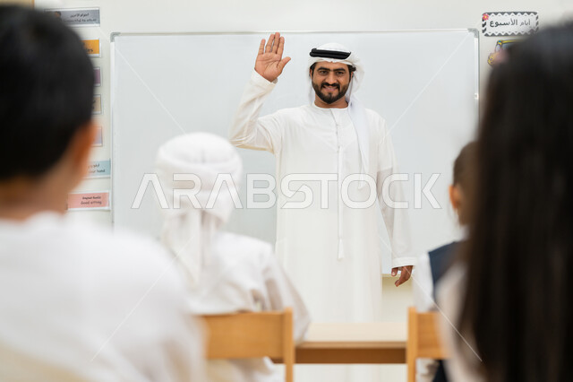 لوحة مدرسية بيضاء، التفاعل والنشاط داخل الفصل الدراسي، المشاركة الفعالة للتلاميذ، معلم عربي خليجي إماراتي يرتدي الكندورة والغترة يرفع كف يده إلى الأعلى للتحية، أهداف التربية والتعليم بالمناهج المقررة للطلاب والطالبات في الإمارات العربية المتحدة