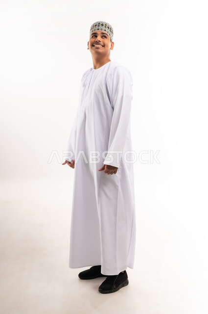 الوقوف باستقامة واعتدال، مفهوم الرجولة والثقة بالنفس، بورتريه لشاب عربي خليجي عماني مبتسم يرتدي الدشداشة و الكمه ينظر الى مكان ما بإيماءات السرور والسعادة، الاهتمام بالأناقة والمظهر الخارجي، صورة الجسم بالطول الكامل, خلفية بيضاء