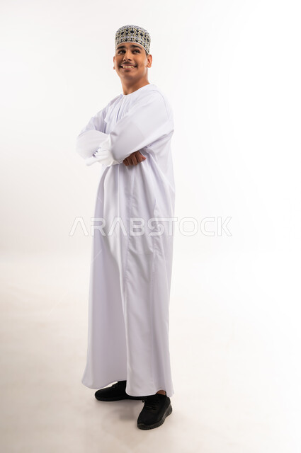 النظر الكاميرا بإيماءات السعادة والسرور, الاهتمام بالأناقة والمظهر الخارجي, تعابير الثقة والاعتزاز بالنفس, بورتريه لرجل عربي خليجي عماني مبتسم يرتدي الدشداشة والكمه يقف باستقامة واعتدال وأيدي متقاطعة, خلفية بيضاء